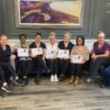 A group of women gather for a photo near a "Beau Institute" backdrop. They are inside a room with a sofa, framed artwork, and a plant. They are holding certificates on their graduation.