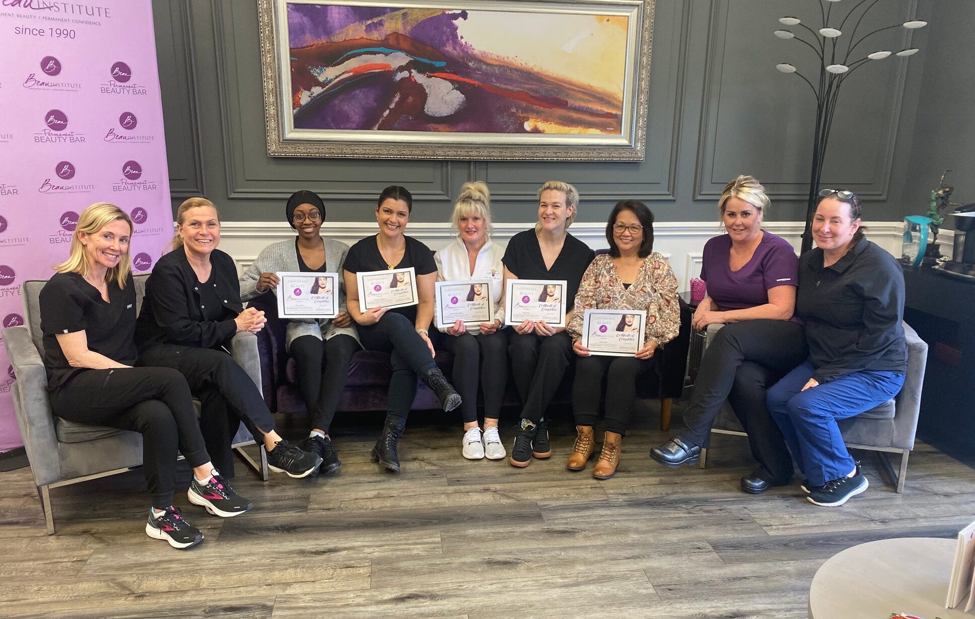 A group of women gather for a photo near a "Beau Institute" backdrop. They are inside a room with a sofa, framed artwork, and a plant. They are holding certificates on their graduation.