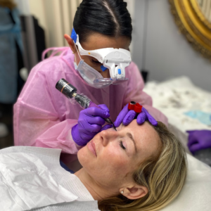 A woman in pink protective gear and masks perform a cosmetic procedure on a patient lying on a table. One of the professionals is using a tool near the patient's eyes.