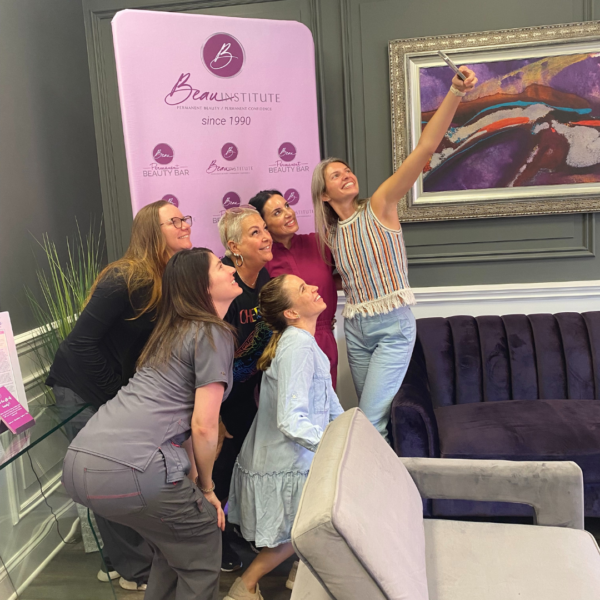 A group of six women gather for a selfie in front of a "Beau Institute" backdrop. They are inside a room with a sofa, framed artwork, and a plant.