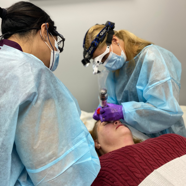 Two people in blue protective gear and masks perform a cosmetic procedure on a patient lying on a table. One of the professionals is using a tool near the patient's eyes.